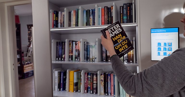 En hand håller upp en bok framför en bokhylla. I hyllorna står flera böcker.