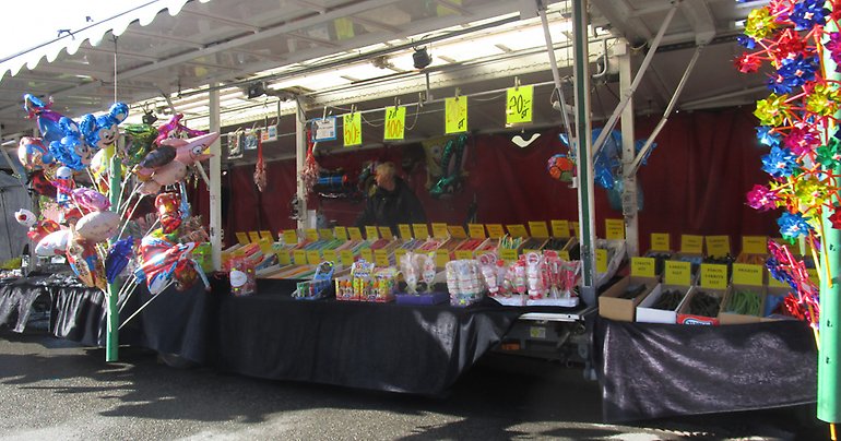 En man står bakom ett marknadsstånd med godis. På vardera sida finns färgglada ballonger.